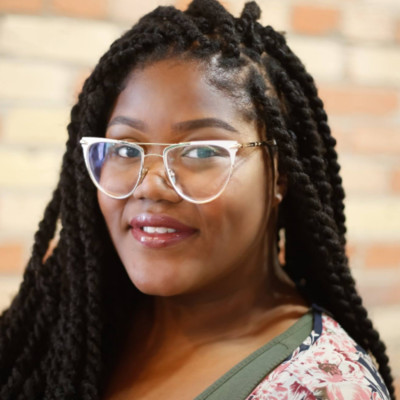 Black woman with clear-rimmed glasses and braided hair smiling with a brick wall behind her.